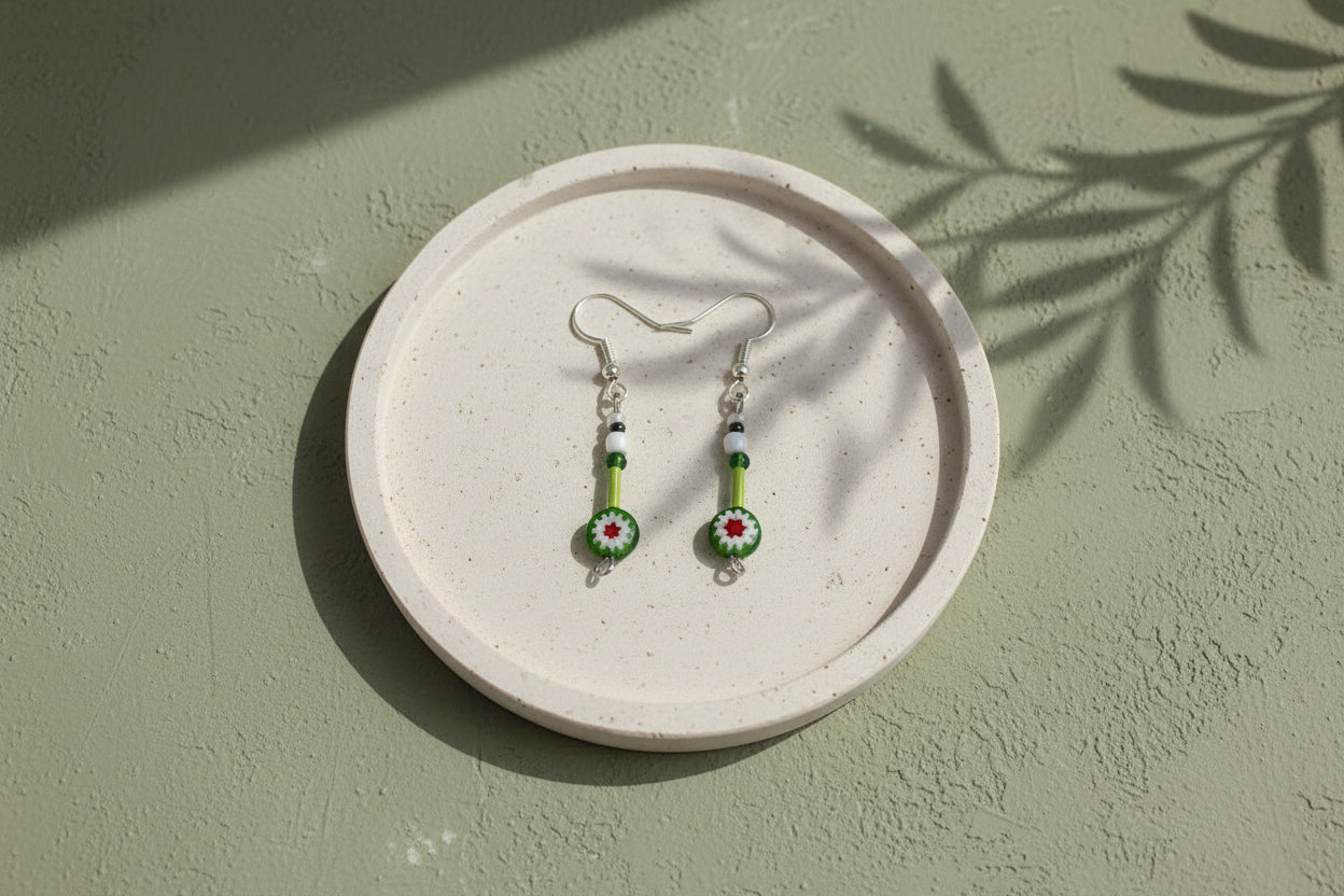 A pair of handcrafted earrings with green and red flower-shaped beads on silver hooks, displayed on a crystal surface.